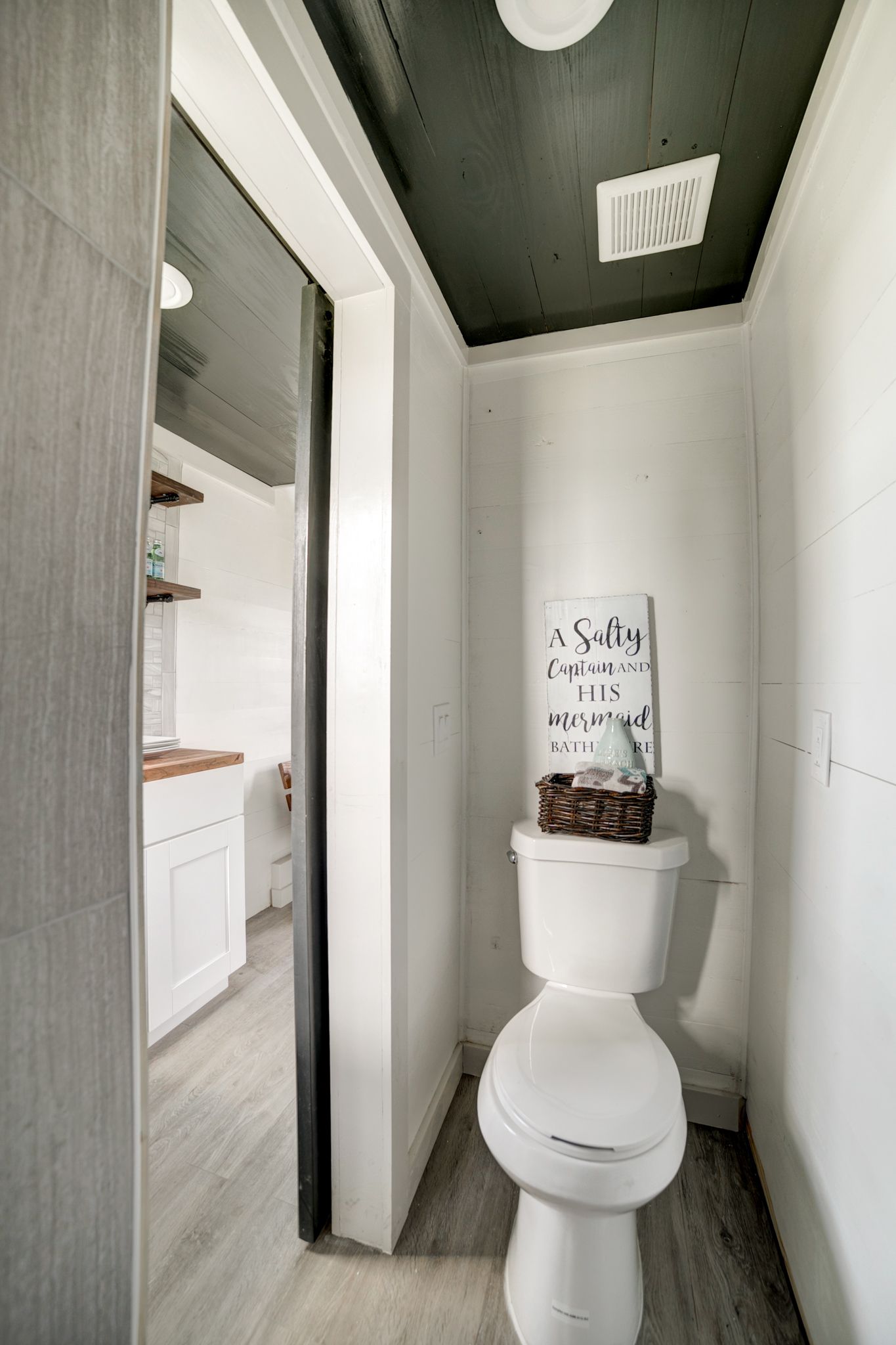 Small tiny house toilet room with white shiplap walls, a dark plank ceiling, a ceiling exhaust fan grille and a sliding door