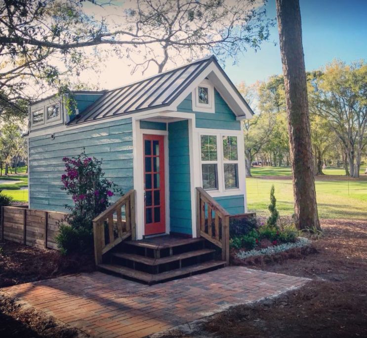Teal tiny cottage with a red front door, metal roof and wooden steps, set among oak trees