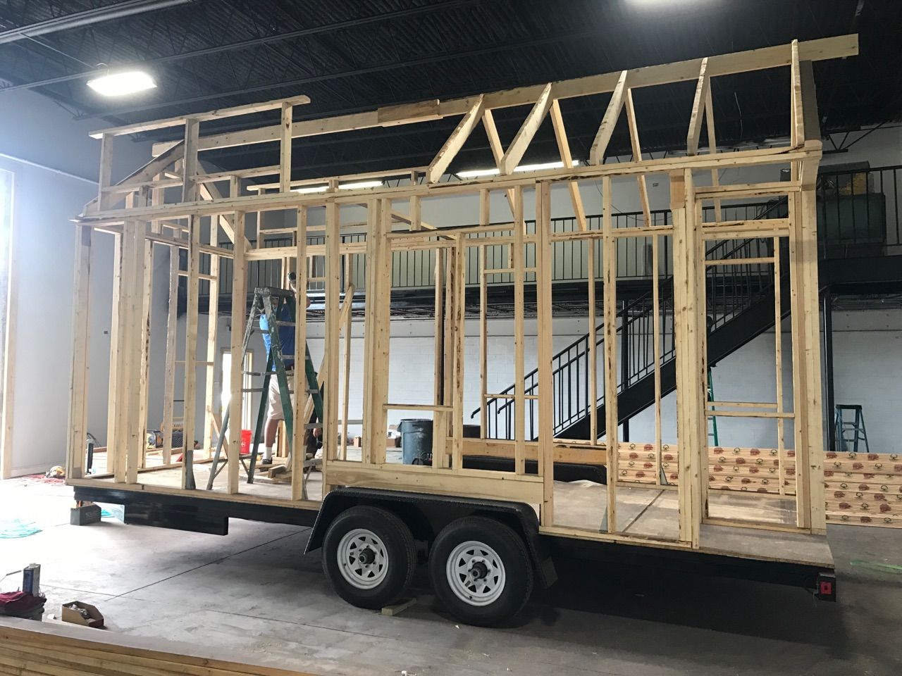 Stud walls and roof framing going up on a 20-foot tandem-axle tiny house trailer inside a workshop
