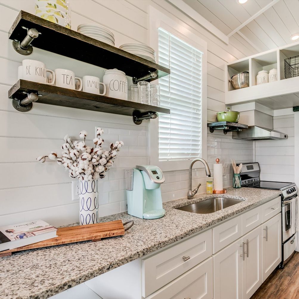 Compact tiny house kitchen with a granite counter, undermount sink, slide-in electric range under a vent hood, and open pipe-bracket shelves