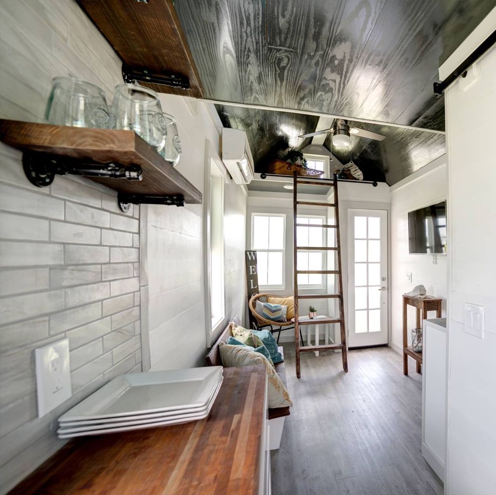 Narrow tiny house interior with a butcher-block counter, open shelves, bench seating and a ladder up to the sleeping loft