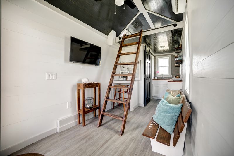 Tiny house living area with a wooden loft ladder, wall-mounted TV and bench seating with pillows