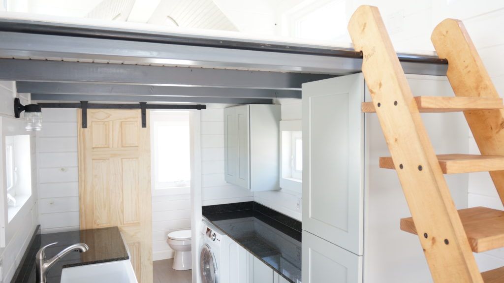 Tiny house galley with tall gray cabinets, a front-load washer under the counter, a barn-door bathroom and a wooden ladder to the loft