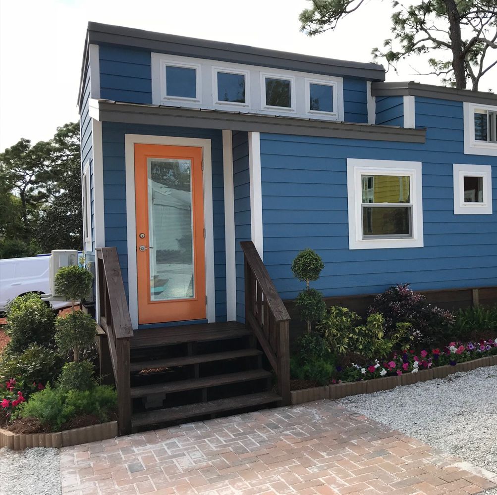 Navy blue tiny house with an orange front door, wooden entry steps and flower beds along the skirting