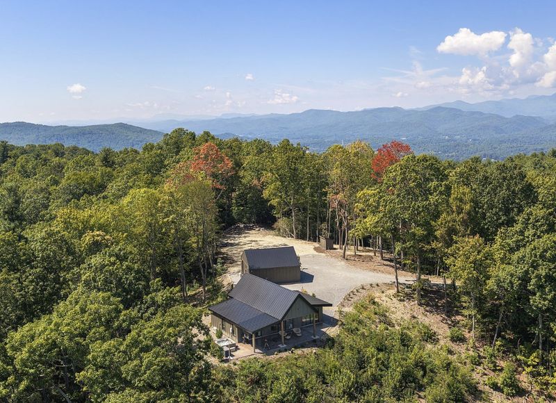 Aerial view of a small dark-roofed house and garage on a wooded ridge with layers of blue mountains behind