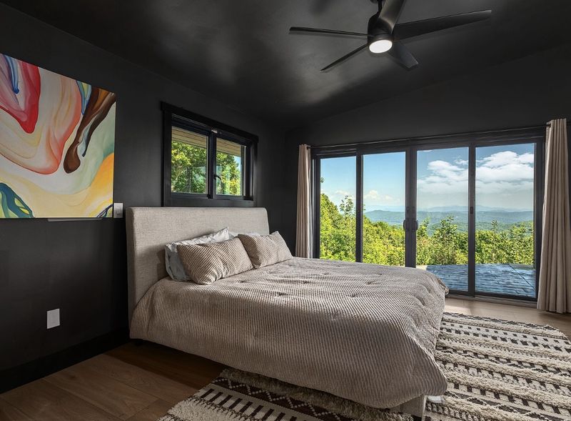 Dark-walled bedroom with a simple upholstered bed, a patterned rug, one abstract painting and a wall of glass doors looking out at mountains