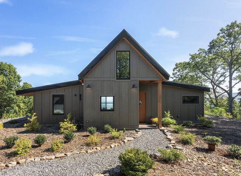 Dark board-and-batten house with a steep gable, black-framed windows, a wood front door and a gravel path through new plantings