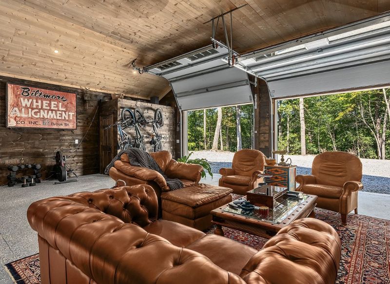 Garage lounge with leather chesterfield sofas, armchairs and a Persian rug, with both roll-up doors open to the woods