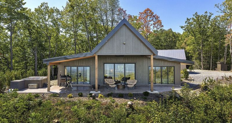 Back of the house with three sets of glass sliding doors opening onto a covered stone patio with a hot tub and grill, surrounded by woods